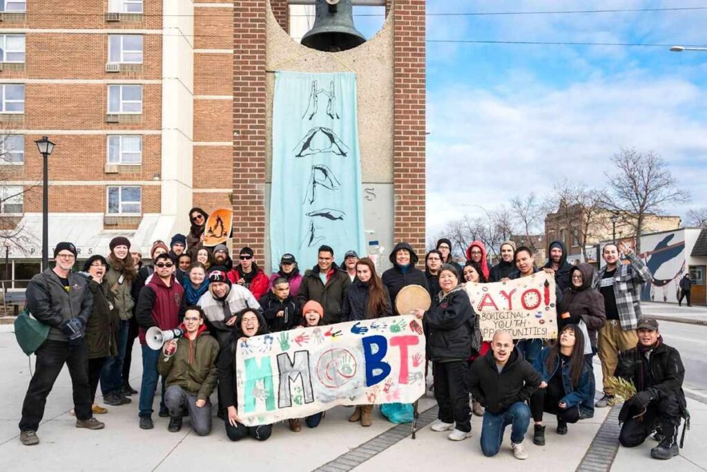 A group of 40 people standing in front of a bell tower and smiling at the camera. A few people are holding handpainted banners, one of which says "AYO! Aboriginal Youth Opportunities!" One person is holding a megaphone. One person is holding a hand drum.