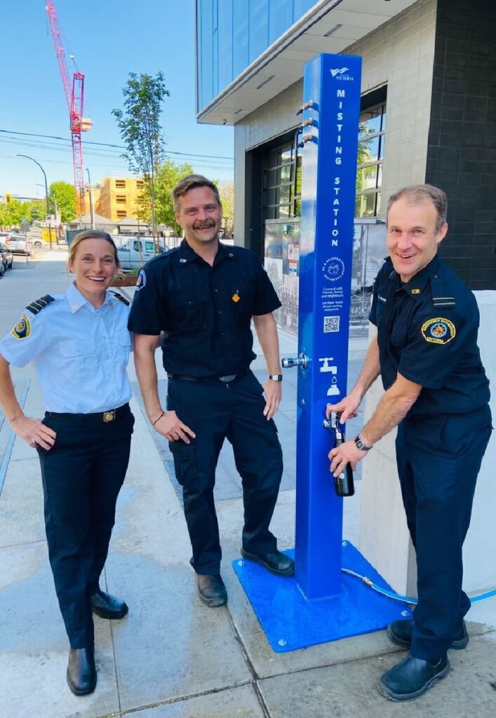 Three emergency management professionals smiling at the camera next to a misting station on a sidewalk. One of the professionals is filling a waterbottle from a spout on the misting station.