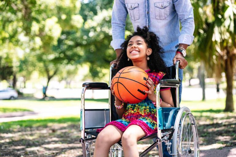 A young girl smiles and holds a basketball. She is sitting in a wheelchair and an adult is holding the back of the chair.